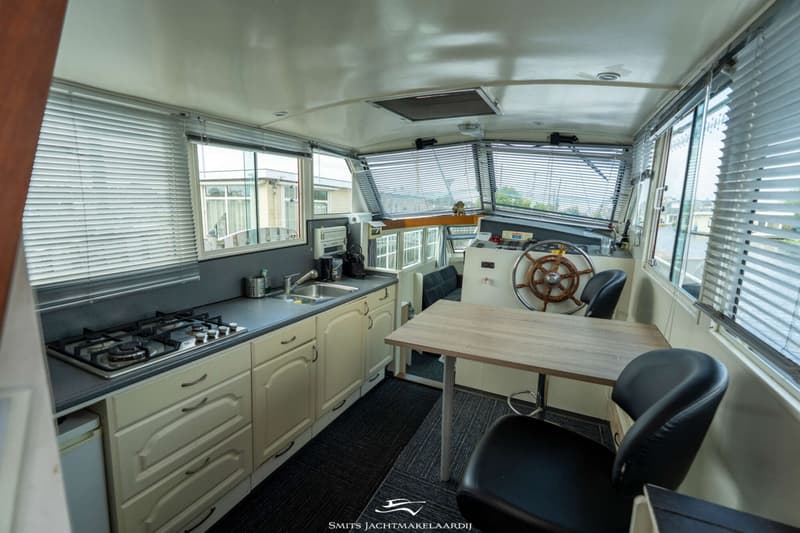 Een interieur van een boot met een kombuis, stuurstand en eethoek, met ramen voorzien van jaloezieën.