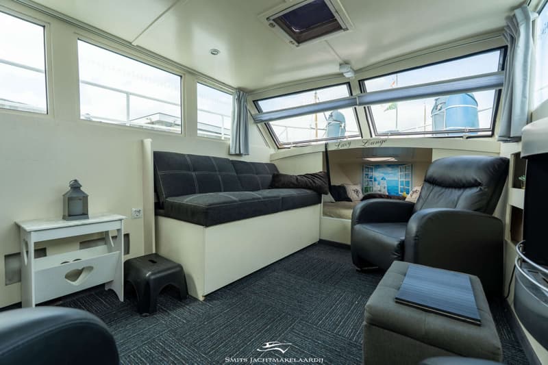De foto toont een interieur van een boot met een bank, fauteuil, poef, tafeltje, en ramen met uitzicht.