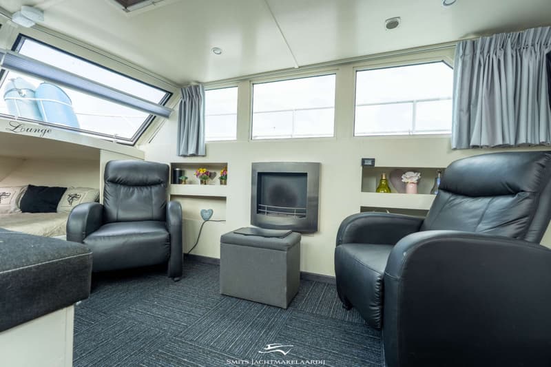Een interieur van een boot met twee zwarte fauteuils, een bed, een open haard en ramen met gordijnen.