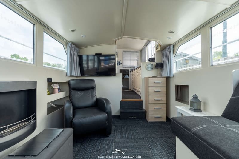 De foto toont het interieur van een boot met een zithoek, een televisie, een doorgang naar een andere ruimte en ramen.