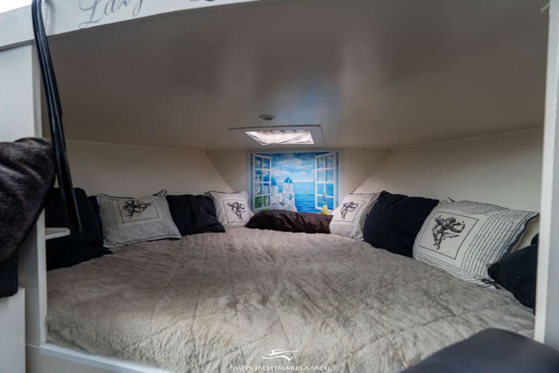 Een kajuit met een tweepersoonsbed, kussens en een muurschildering van een Grieks landschap met de zee.