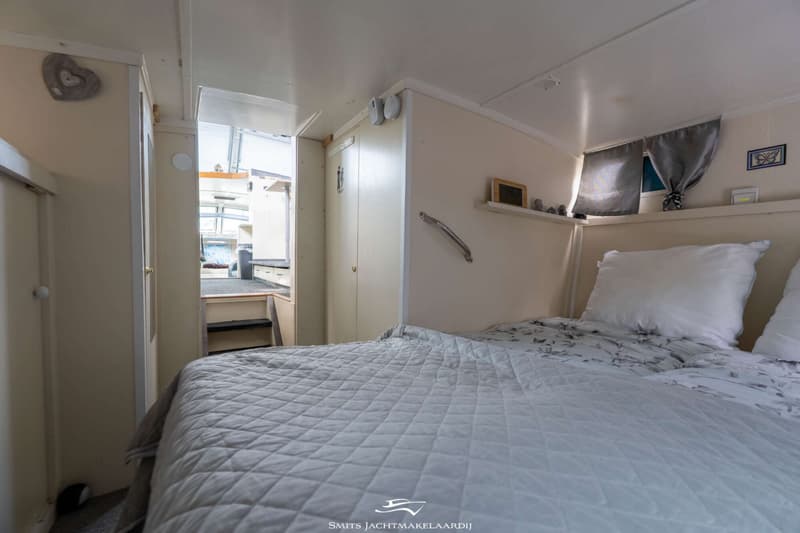 Een slaapkamer aan boord met een opgemaakt bed, kasten, een raam met gordijnen en een doorgang naar een andere ruimte.