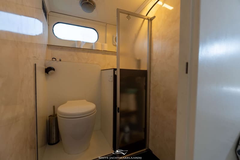 Een badkamer met een wit toilet, een toiletborstel, een patrijspoort, en een douchecabine met een glazen deur.