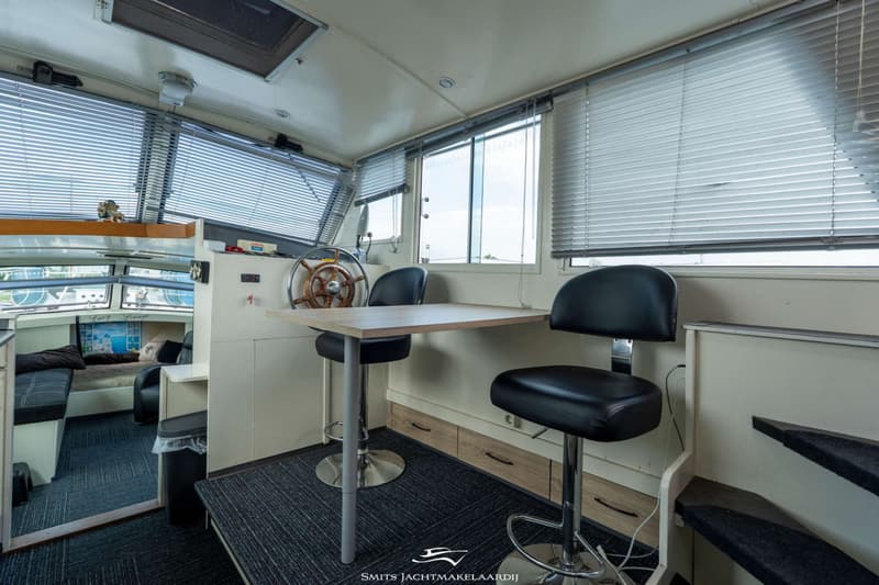De foto toont een interieur van een boot met een stuurwiel, een tafel met twee barkrukken, en jaloezieën voor de ramen.