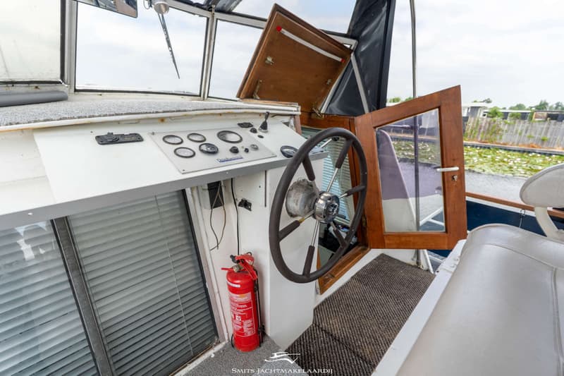 De foto toont de stuurstand van een boot met een stuurwiel, instrumentenpaneel, brandblusser en een open houten deur.