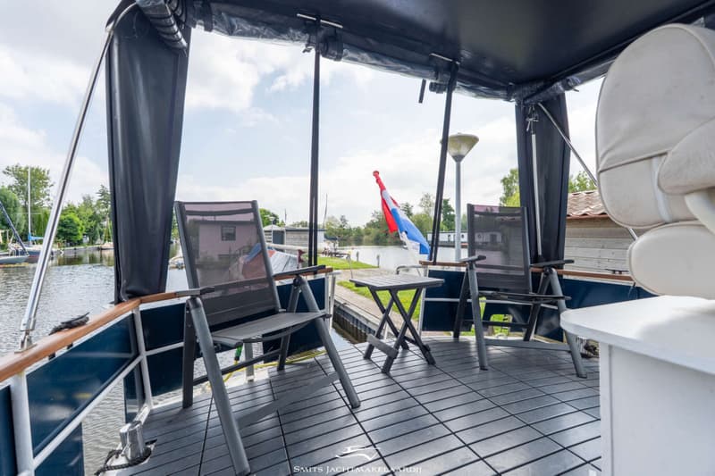 Op het achterdek van een boot staan twee grijze klapstoelen en een klein klaptafeltje onder een donkerblauwe bimini.