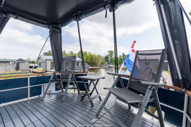 Een dek met twee klapstoelen en een klaptafel onder een donkerblauwe bimini, met een Nederlandse vlag aan stuurboord.