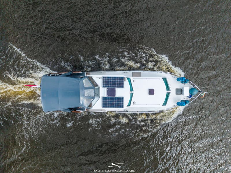 Een witte motorboot met een grijs dekzeil en zonnepanelen vaart over donker water, gezien van bovenaf.