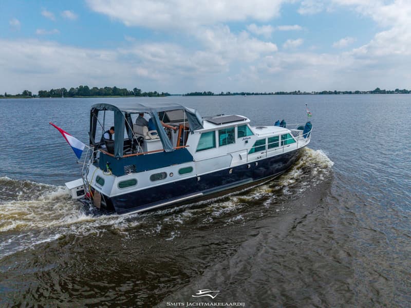 Een blauw-witte motorboot met de naam "Jottum" vaart op een meer onder een bewolkte hemel, met een Nederlandse vlag aan de…