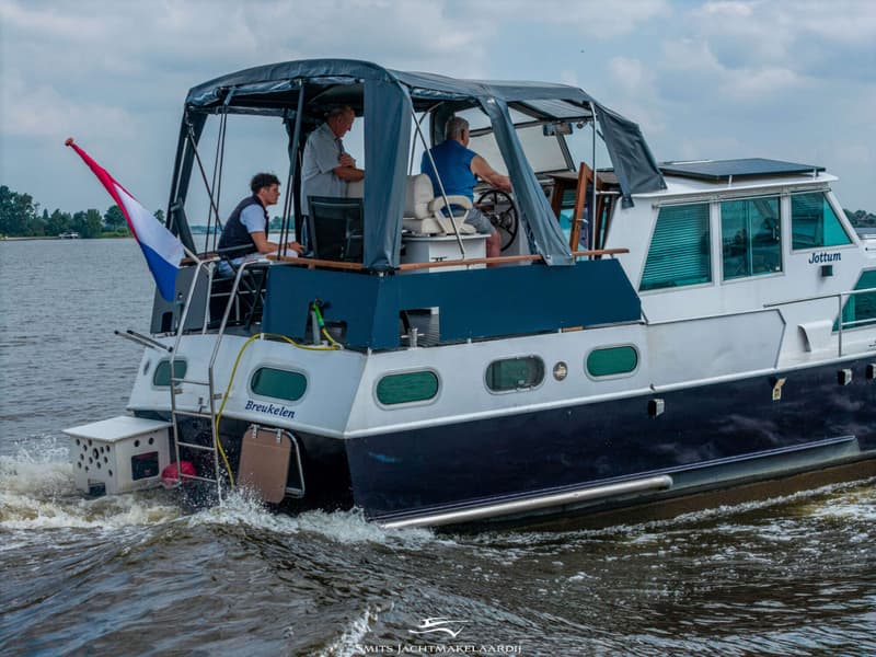 Een blauw-witte motorboot met drie personen aan boord vaart op water, met de Nederlandse vlag aan de achtersteven.