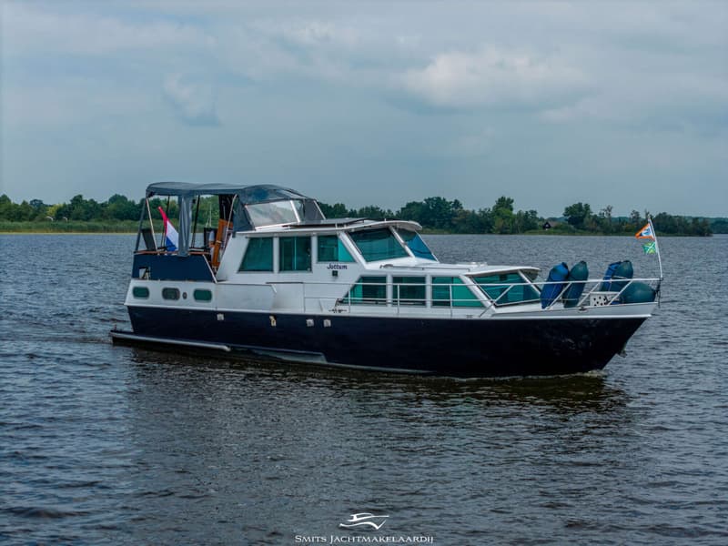 Een donkerblauwe en witte motorboot met de naam "Jottum" vaart op een meer met bomen op de achtergrond.