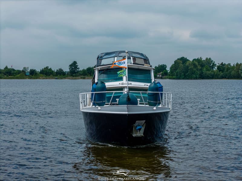 Een donkerblauwe boot met een witte opbouw en een oranje vlag met "EOC" erop vaart op een meer onder een bewolkte lucht.