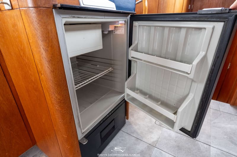 Een openstaande, lege koelkast met een klein vriesvak en een rooster, ingebouwd in een houten interieur.