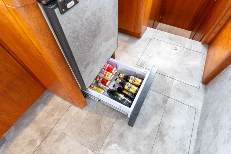 Een open koellade met blikjes frisdrank, bierflesjes en een fles mousserende wijn, omringd door houten panelen en een…