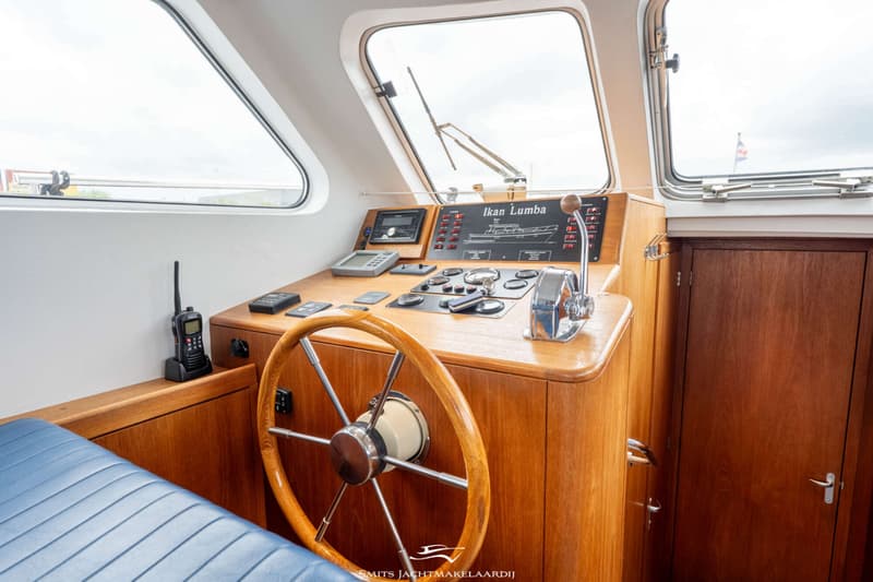 De foto toont het stuurhuis van een boot met een houten stuurwiel, diverse navigatie-instrumenten en een blauwe bank.