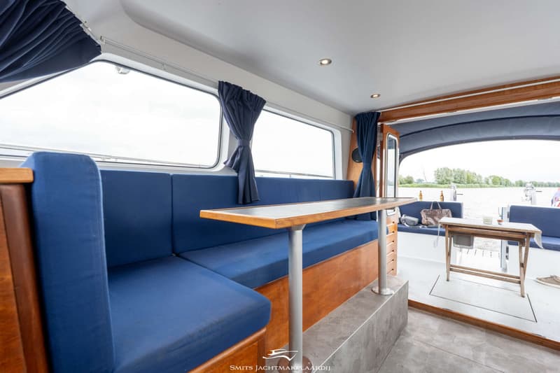Een interieur van een boot met blauwe banken, een houten tafel en uitzicht op water met bomen.