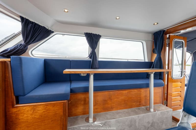 De foto toont een interieur van een boot met blauwe banken, een houten tafel en ramen met blauwe gordijnen.