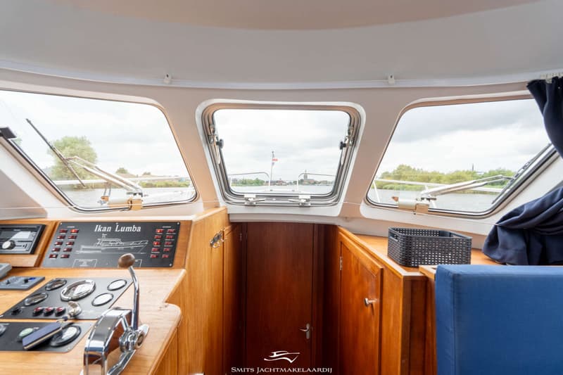 De foto toont het interieur van een boot met een stuurstand, houten afwerking, ramen en uitzicht op water en land.