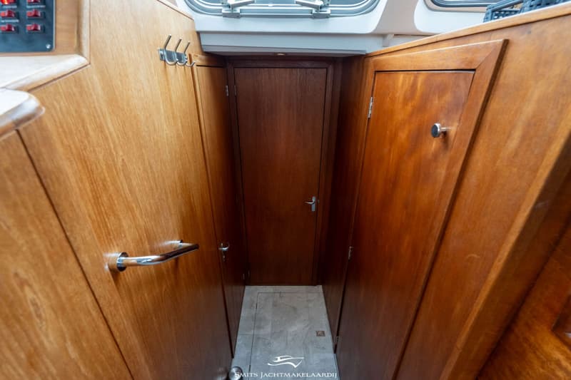 Een smalle gang met houten wanden en deuren, een grijze vloer en een raam bovenin.
