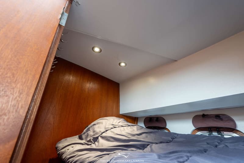 Een kajuit met een bed met grijs dekbed, houten wanden, twee patrijspoorten en twee plafondlampen.