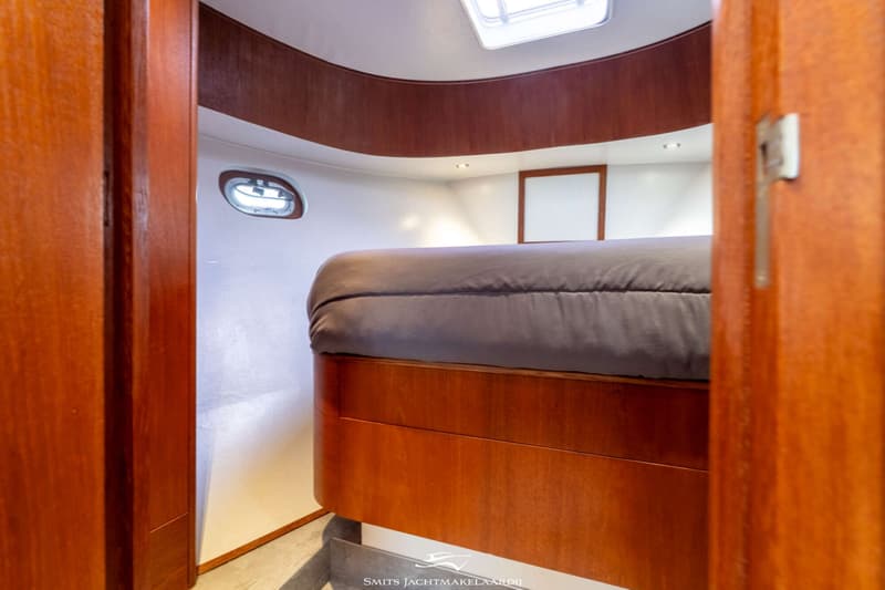 Een kajuit met een bed met donkergrijze hoes, houten afwerking, een patrijspoort en een dakluik.
