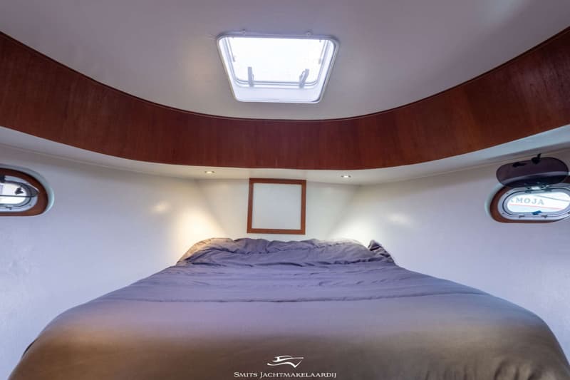 Een kajuit met een bed, twee patrijspoorten, een dakluik en een houten omlijsting aan het plafond.