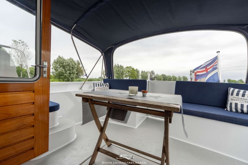 Een houten klaptafel staat op het dek van een boot met blauwe banken en een vlag op de achtergrond.