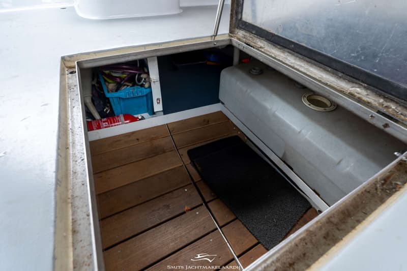 Een open luik in de vloer van een boot toont een brandblusser, een blauwe krat, een grijze tank en een houten vloer.