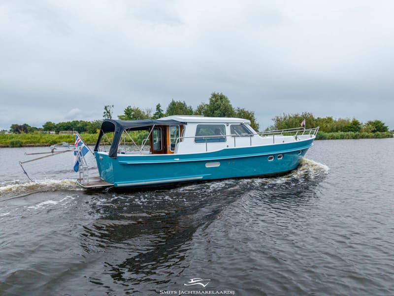 Een blauw-witte motorboot vaart op een waterweg met bomen en struiken op de achtergrond onder een bewolkte lucht.