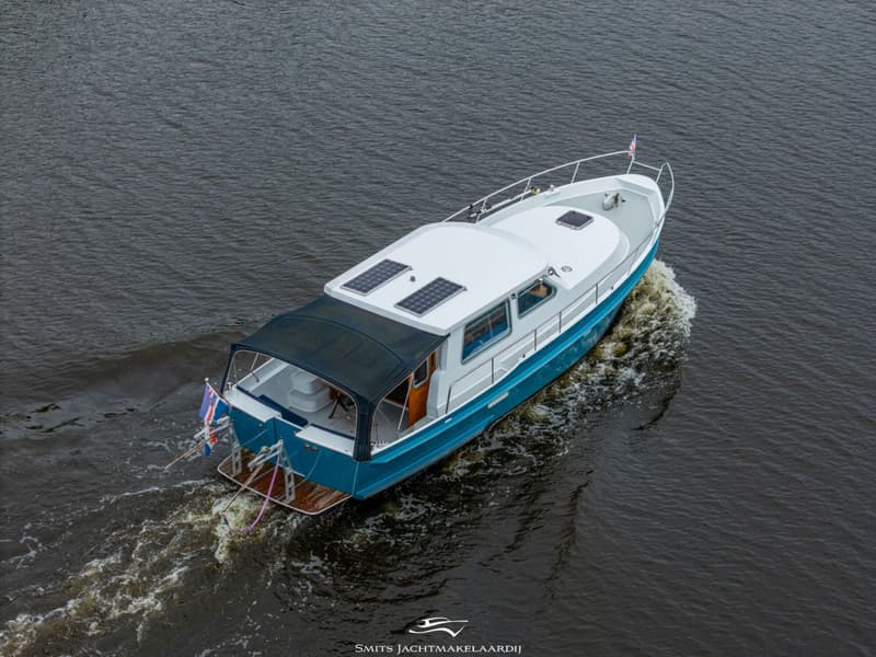 Een blauw-witte motorboot met zonnepanelen op het dak vaart over donker water, gezien van bovenaf.