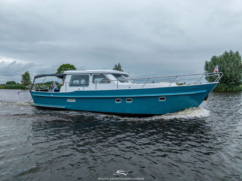 Een blauw-witte motorboot vaart op het water, met een Nederlandse vlag aan de boeg en een logo van "SMITS JACHTMAKELAARDIJ"…