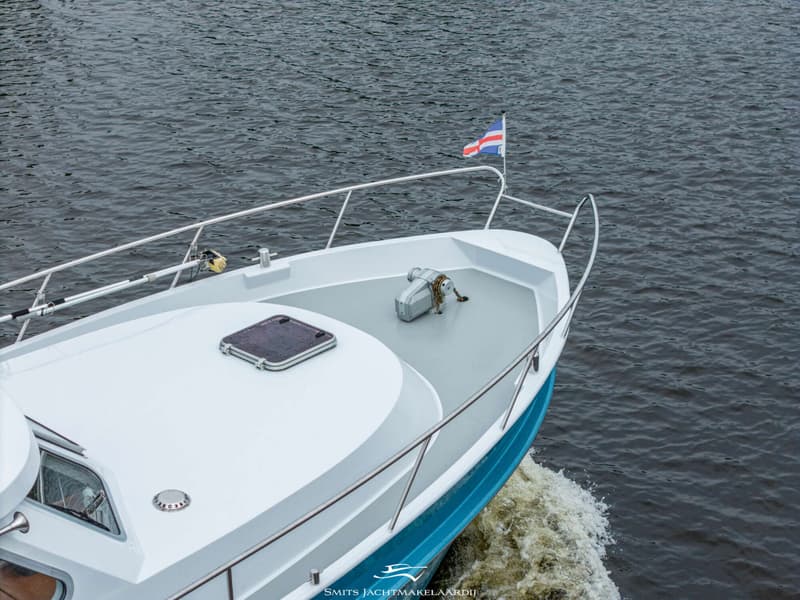 Een blauw-witte boot met een vlag op de boeg vaart over donker water, met een ankerlier en reling zichtbaar.