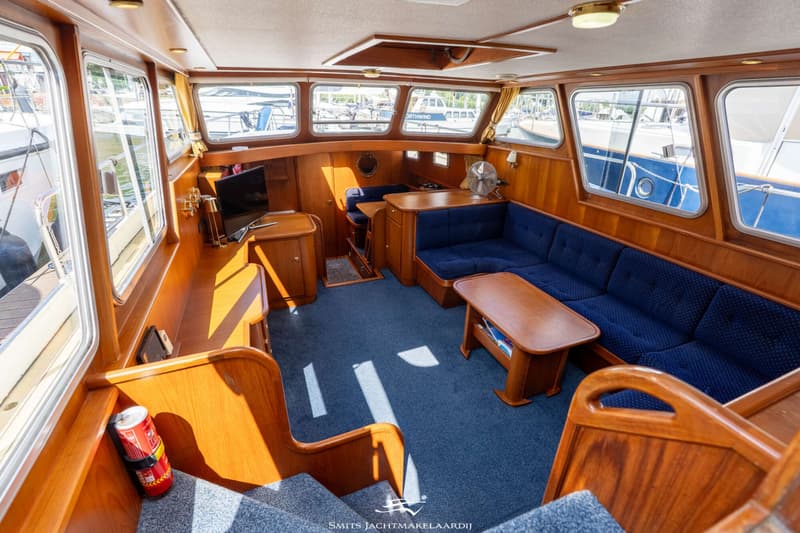 De foto toont het interieur van een boot met houten afwerking, blauwe bekleding en tapijt, en ramen rondom.
