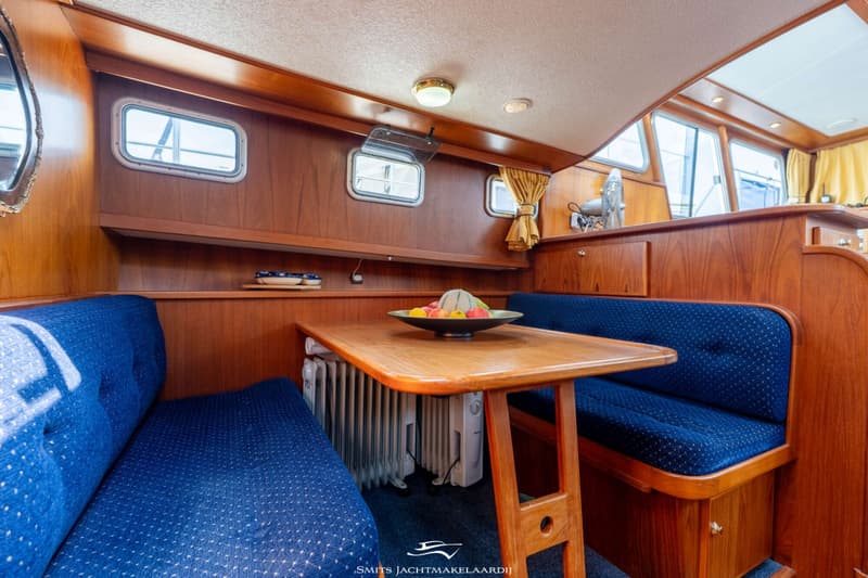 De foto toont een houten interieur van een boot met blauwe banken en een tafel met een fruitschaal.