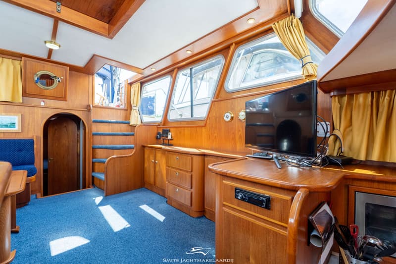 De foto toont het interieur van een boot met houten wanden, een blauw tapijt, ramen met gele gordijnen en een televisie.