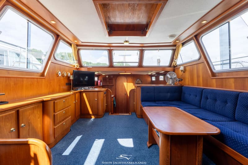 De foto toont het interieur van een boot met houten lambrisering, blauwe vloerbedekking, een blauwe bank en een houten tafel.