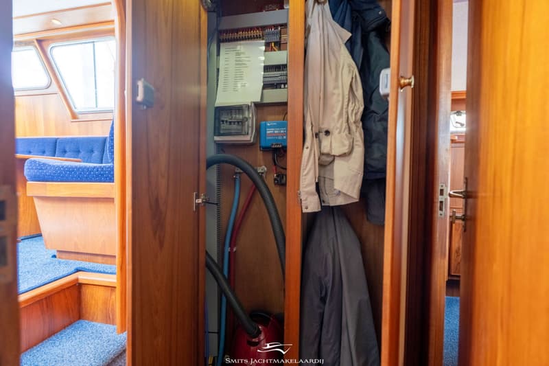 Een open kast met kleding, een stofzuiger, elektrische panelen en slangen, met op de achtergrond een blauwe bank en ramen.
