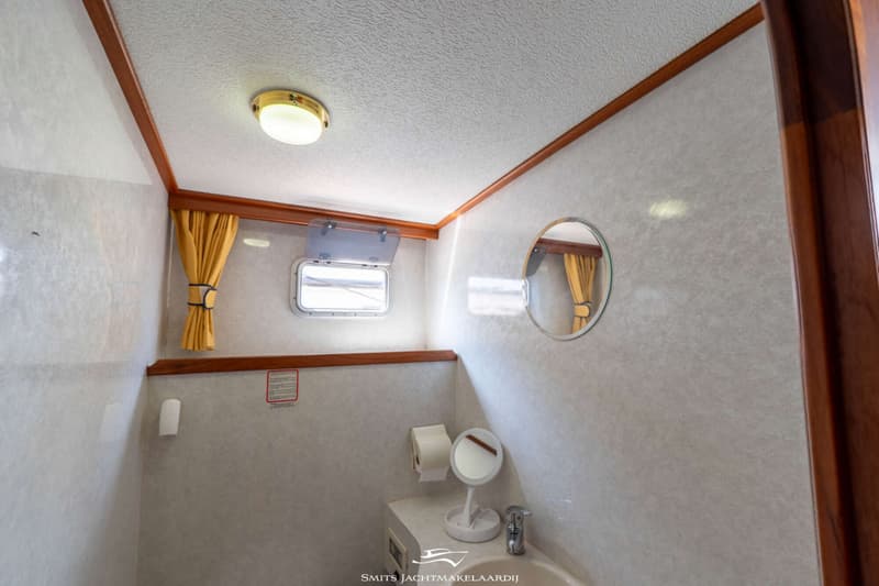 Een interieur van een boot met een plafondlamp, een open raam, een spiegel, een toiletrolhouder en een wastafel met kraan.