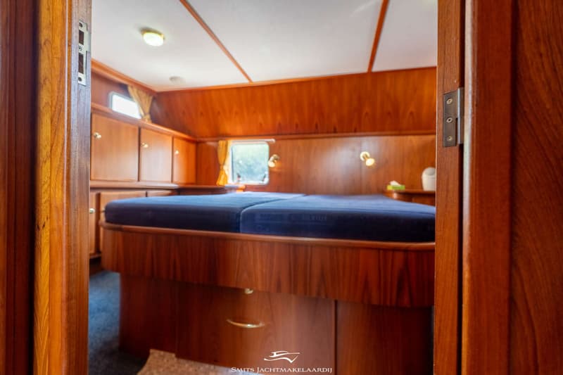 Een houten kajuit met een tweepersoonsbed, kasten, een raam met gordijnen en twee leeslampjes.
