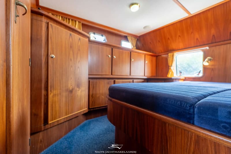 Een kajuit met een tweepersoonsbed, houten kasten en panelen, een raam met gordijnen en een patrijspoort.