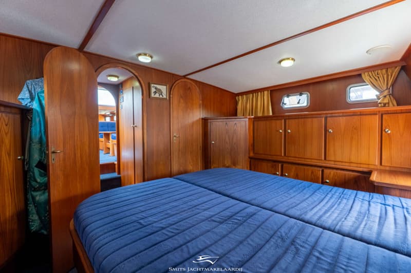 Een slaapkamer in een boot met een groot blauw bed, houten wanden en kasten, en twee ramen met gordijnen.