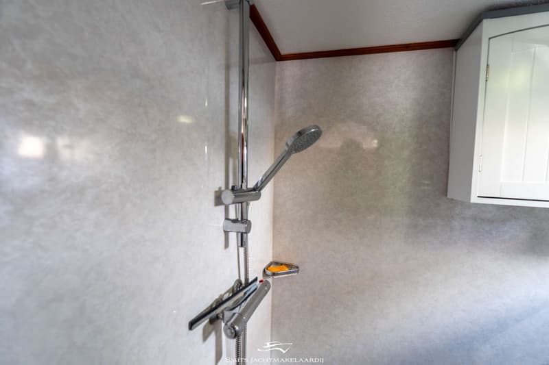Een chromen douchekop en stang zijn gemonteerd aan een lichtgrijze wand, met een stuk zeep in een houder.