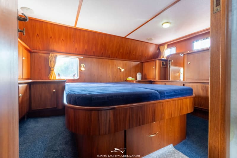 Een slaapkamer in een boot met een groot bed, houten wanden en kasten, en een blauw tapijt.