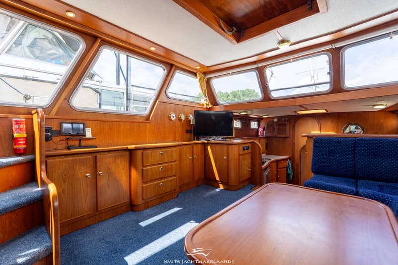 De foto toont het interieur van een boot met houten lambrisering, blauwe vloerbedekking, een tafel, banken en een televisie.