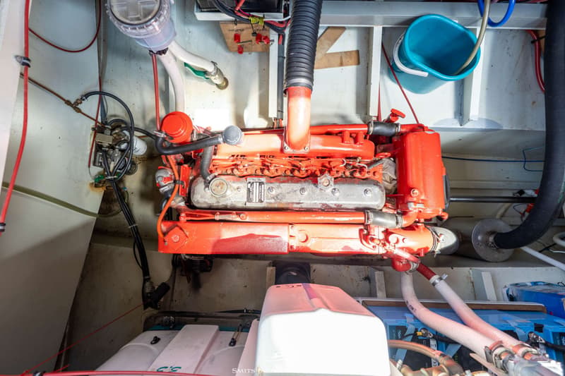 Een rode scheepsmotor met grijze kleppendeksel is zichtbaar in een motorruimte met diverse slangen en bedrading.