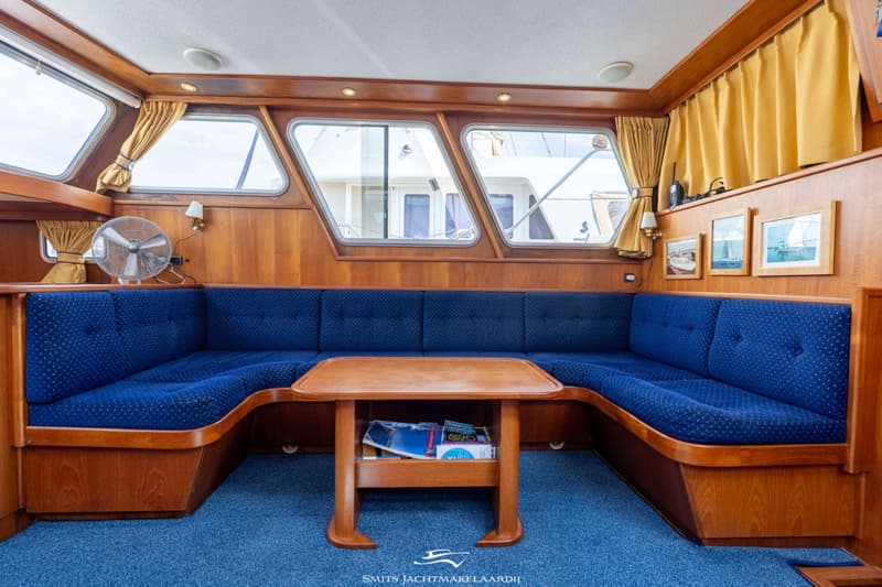 Een interieur van een boot met een blauwe bank, houten tafel, houten wanden en ramen met gele gordijnen.