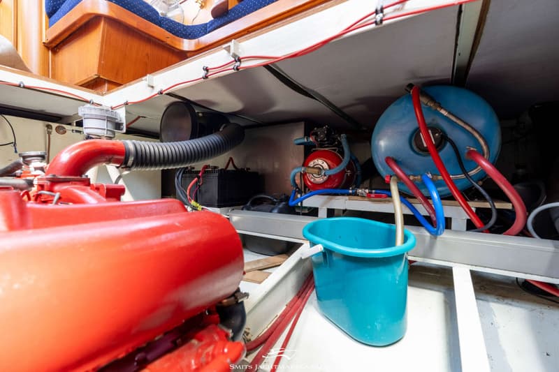 Een rode motor, twee blauwe tanks, een zwarte accu en een blauwe emmer zijn zichtbaar in de motorruimte van een boot.