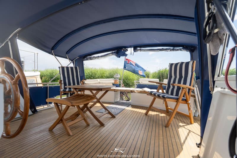 De foto toont een houten dek met een blauwe bimini, houten meubels en een vlag met de Union Jack.
