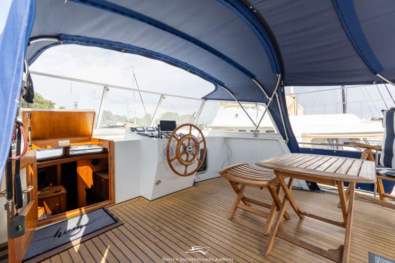 Een houten stuurwiel en instrumentenpaneel zijn zichtbaar op een bootdek met een blauwe bimini en houten vloer.