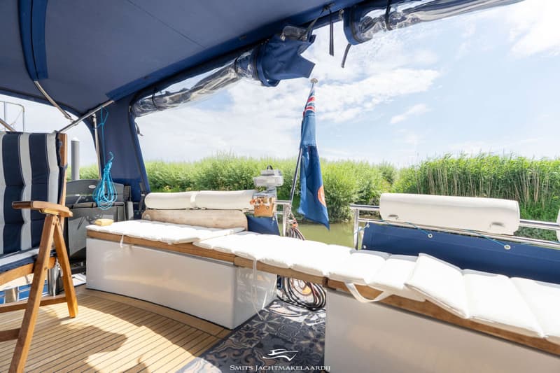 Een bootdek met teakhouten vloer, blauwe bimini, witte kussens op banken, een stoel en een vlag aan de achterzijde.